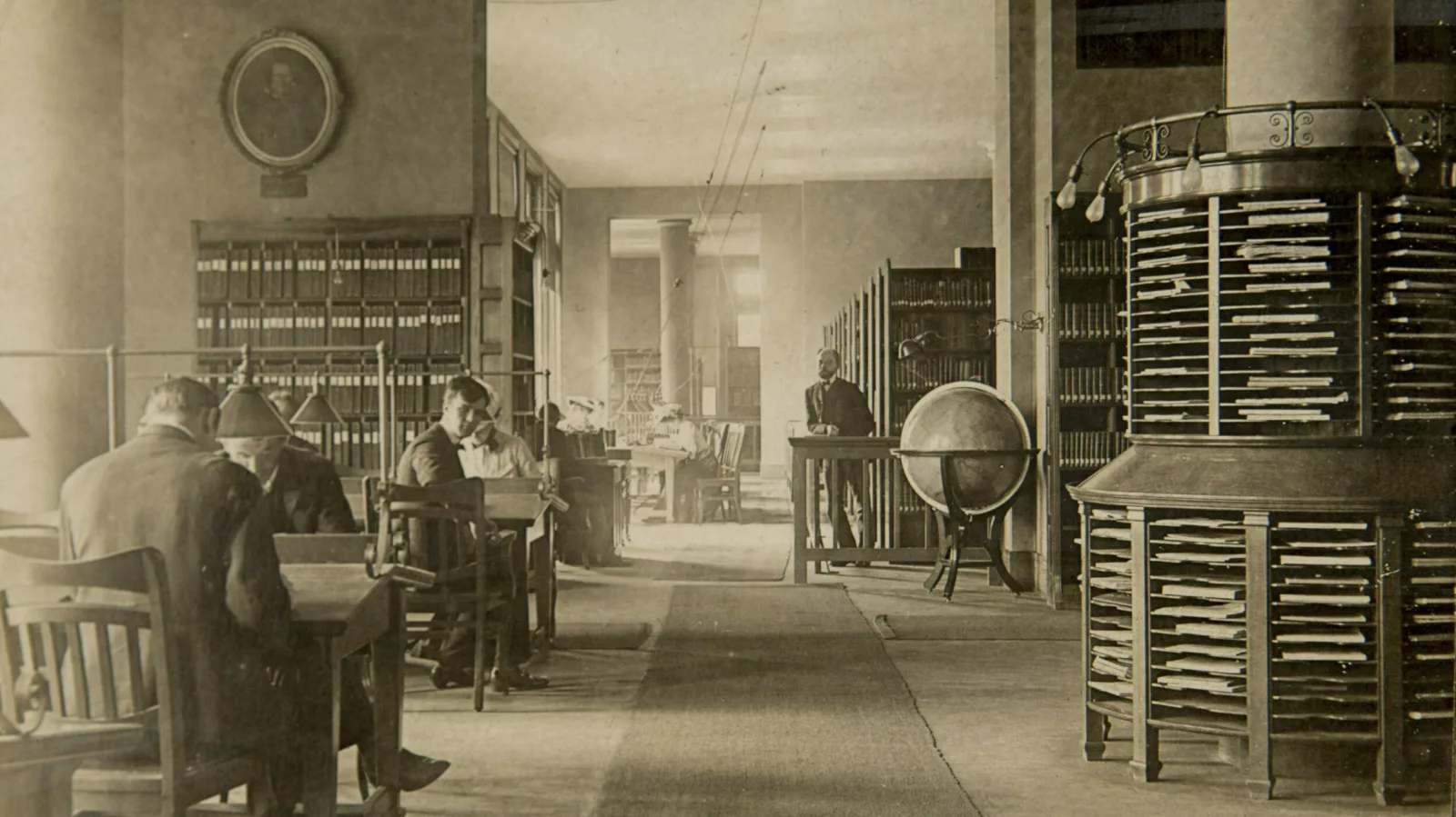 An old photo from around 1900 shows several people studying at desks. One person looks over his right shoulder at the photographer.