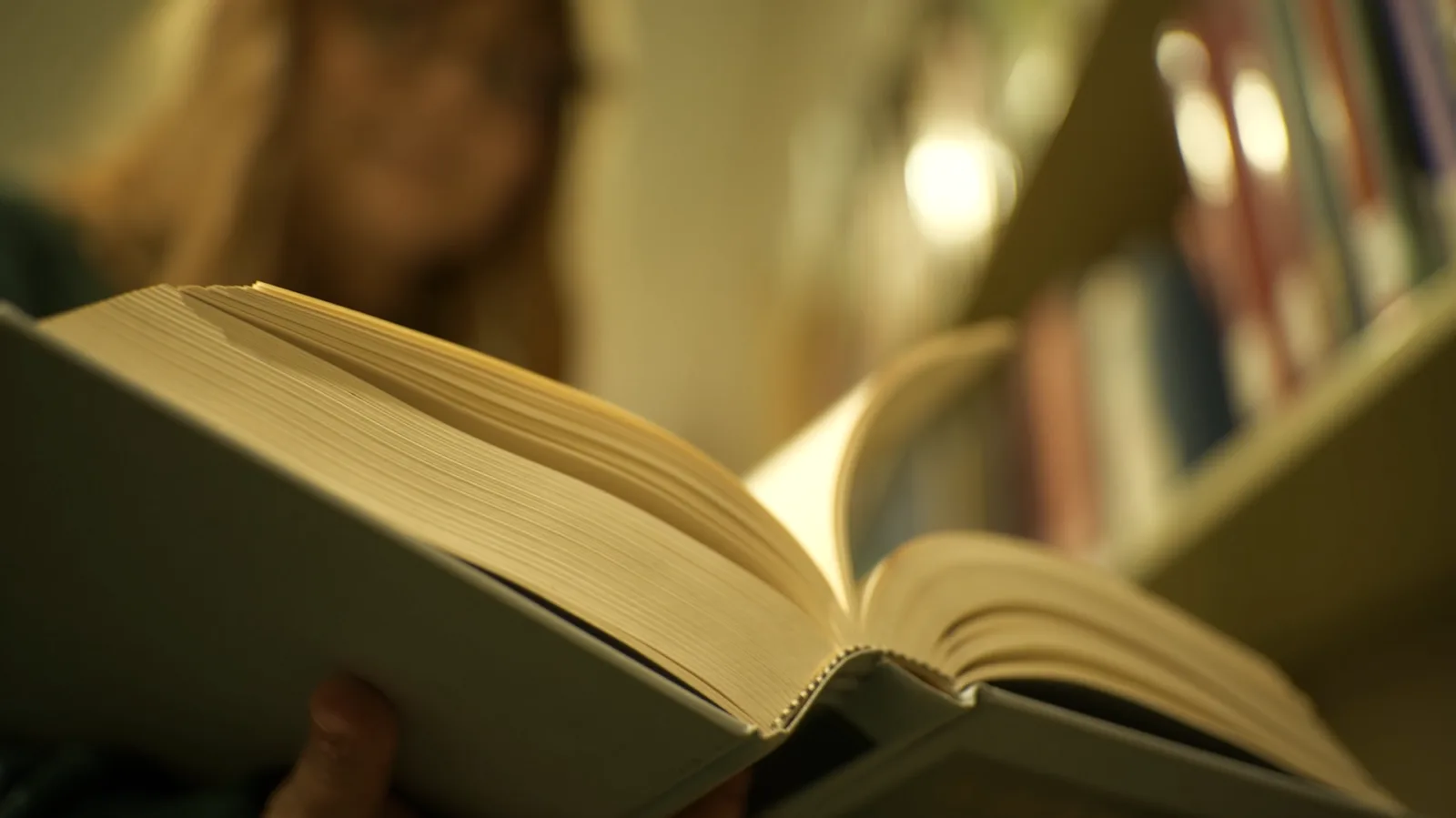 Someone holds a book open and reads from the book. Behind them are shelves of books.