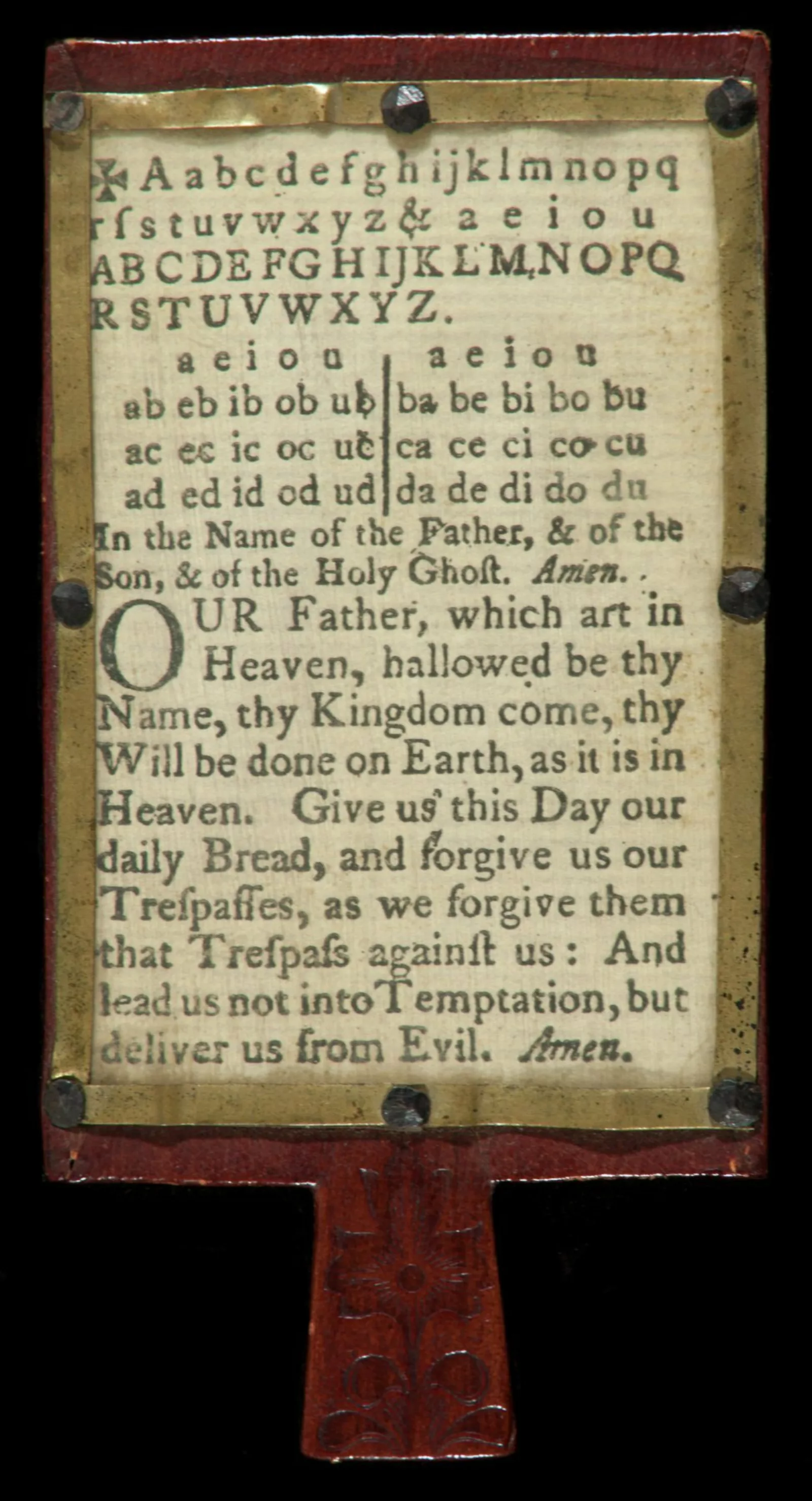 A metal frame is mounted to a wooden board. Within the frame is a printed sheet of paper with the alphabet and the lord’s prayer on it.