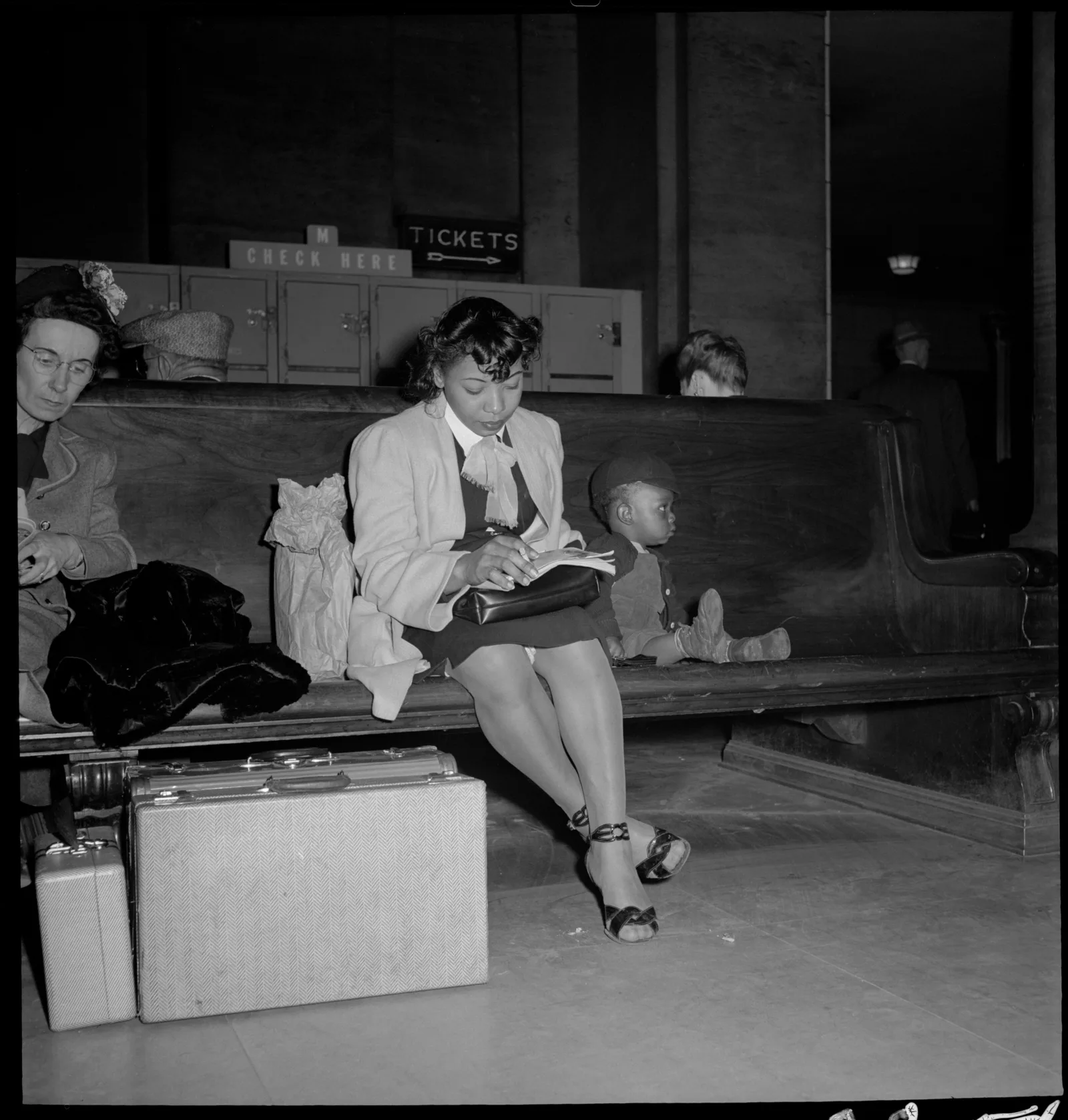 A woman and young boy sitting on a bench in the waiting room, luggage at their feet. The woman is reading, the boy is gazing off to his left.
