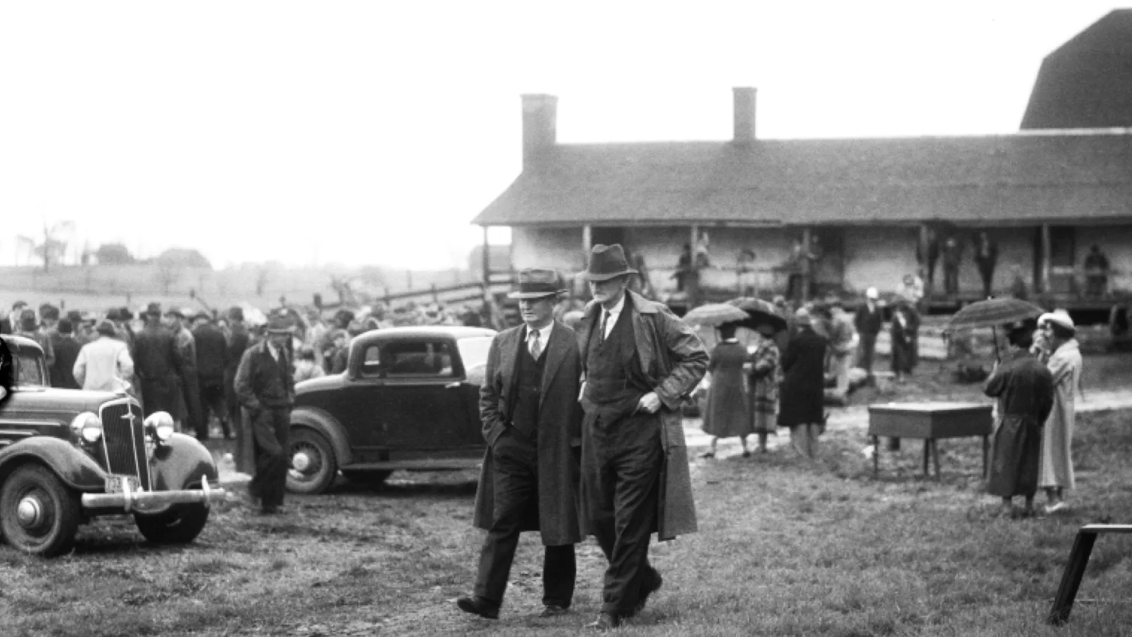 Two men walking through lot filled with cars and people, buildings in background