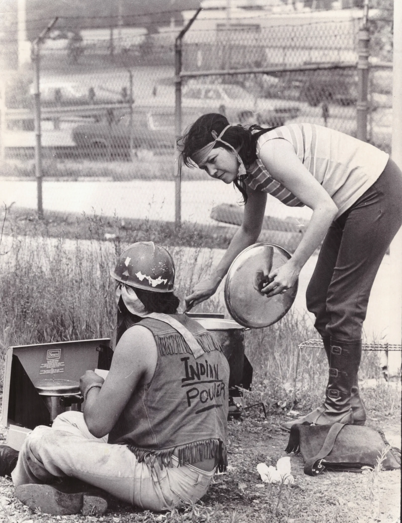 Carol Warrington Warrington is wearing a striped top, black pants, and headband. She is holding a lid and leaning over a pot. To her left is another protestor wearing a helmet and vest that reads "Indian Power."