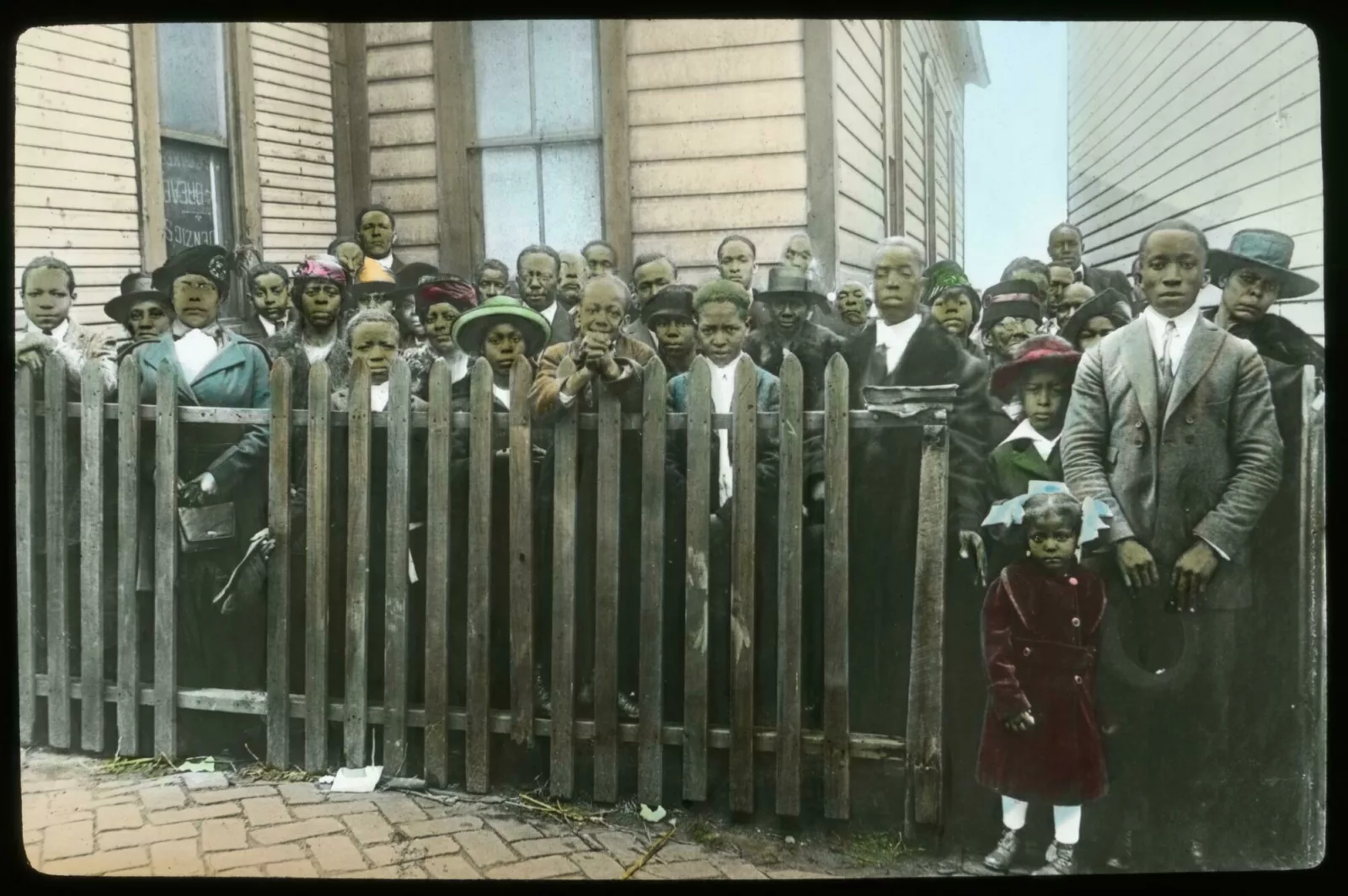 A multi-generational group of people pose for a photo. Some of them are standing behind a wooden picket fence between two buildings.
