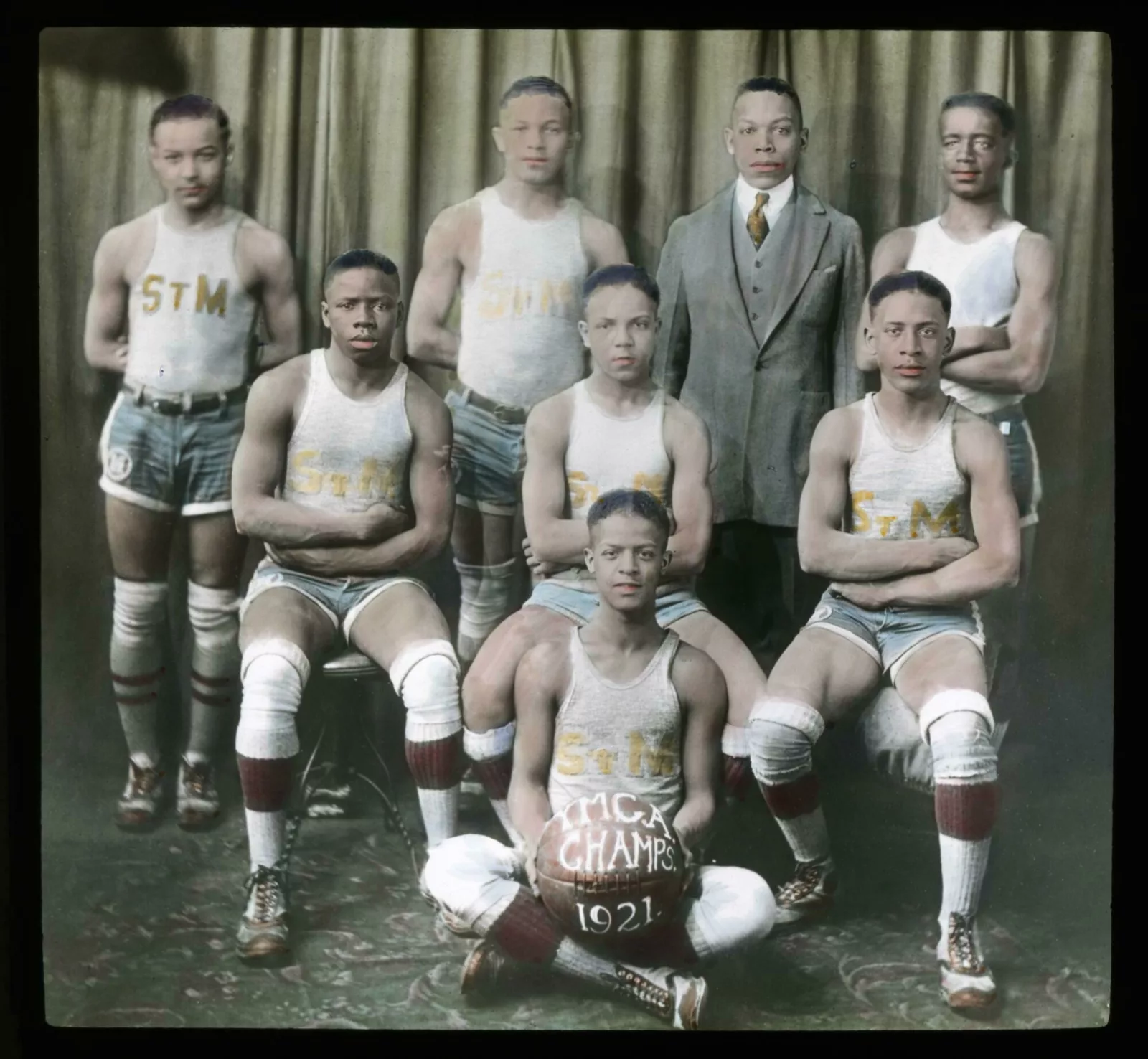 Youth basketball players sit or stand in three rows. They are wearing tank tops, shorts, long socks, and knee pads. The player sitting in the middle holds a basketball. On the basketball are the words “YMCA Champs 1921.”