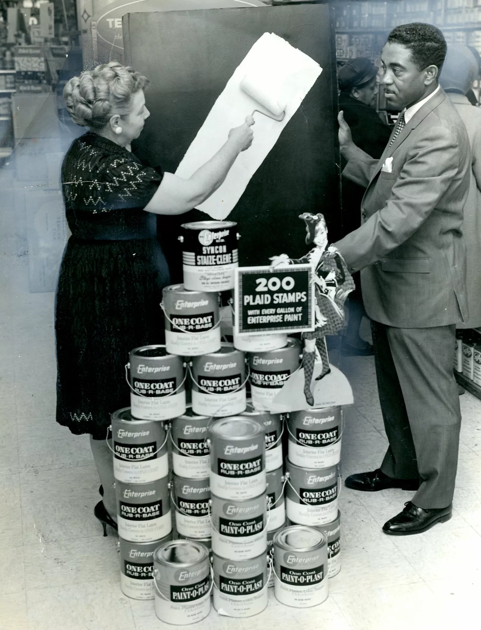 Eunice demonstrating one coat paint at Ebony Paint with store manager Harold Perkins. They are standing behind a display of paint cans.