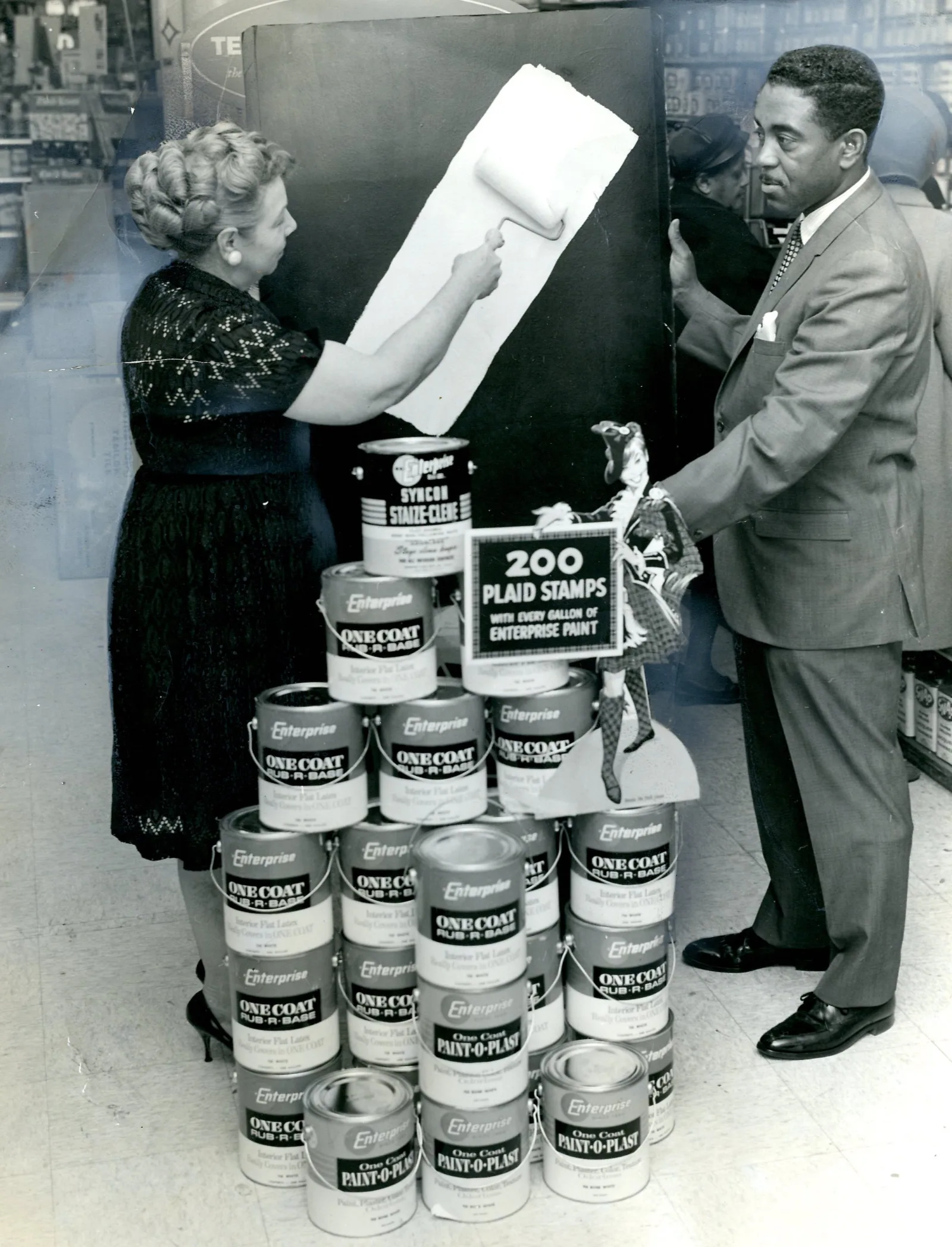 MMS Prescott 044 Eunice Prescott demonstrating one coat paint Eunice demonstrating one coat paint at Ebony Paint with store manager Harold Perkins. They are standing behind a display of paint cans.