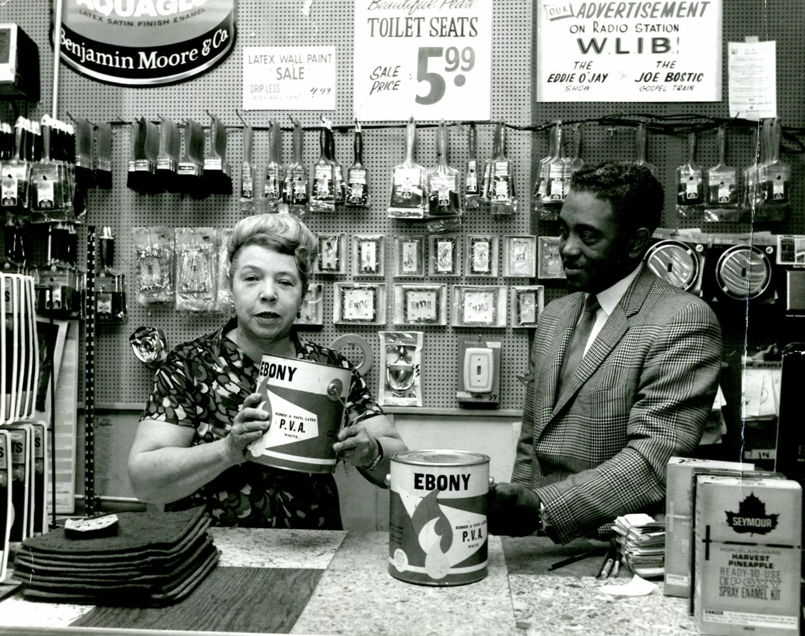 Eunice Prescott holding a can of her paint in her store. On the right is Harold Perkins listening to Eunice talk.