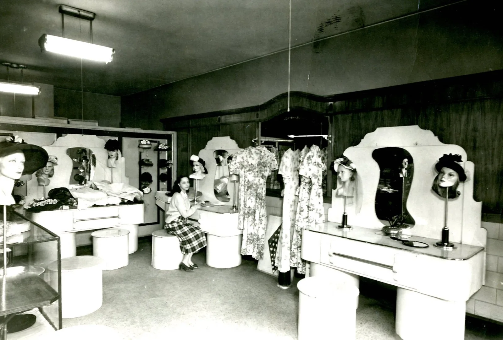 A girl sits on a stool surrounded by mannequin heads wearing hats