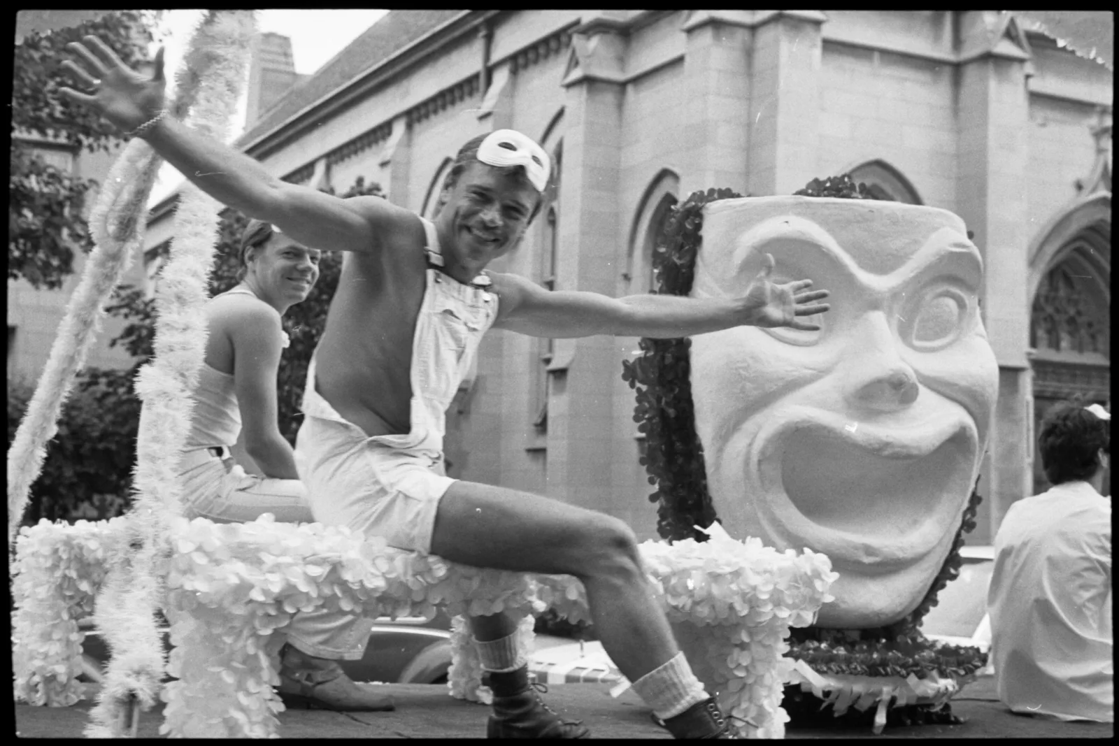 Two men dressed in white sit on a float in the parade. The float has a big mask on it.