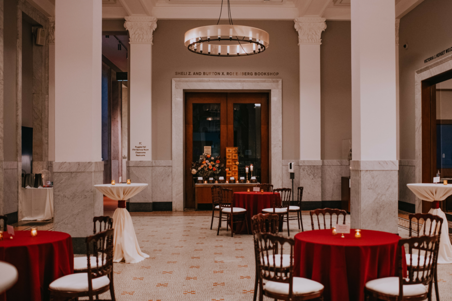 The Newberry lobby set up for a cocktail party. There are tables with red tablecloths and standing cocktail tables with white tablecloths throughout the lobby. In the background, under the chandelier, is the entrance to the Newberry bookshop.